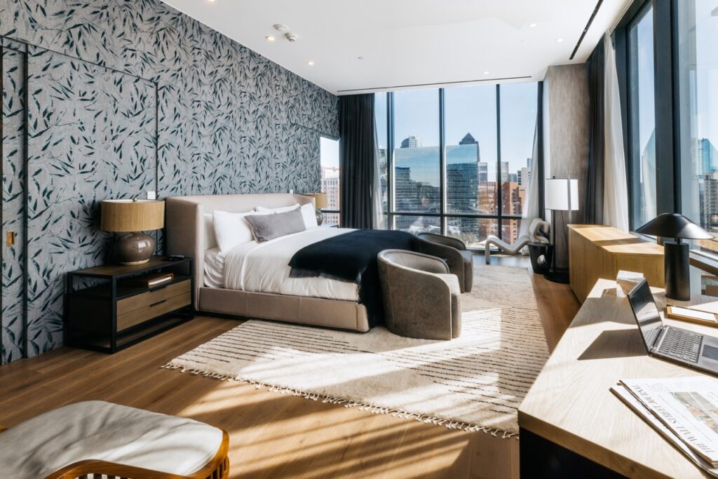 The inside of a hotel room in Dallas with a big bed, large window looking into the city, and a brown wood floor. 
