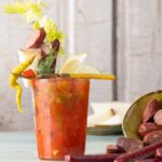 A bloody mary sits in a glass on a table beside New Mexico sausages on the table and veggie garnishes on the drink.