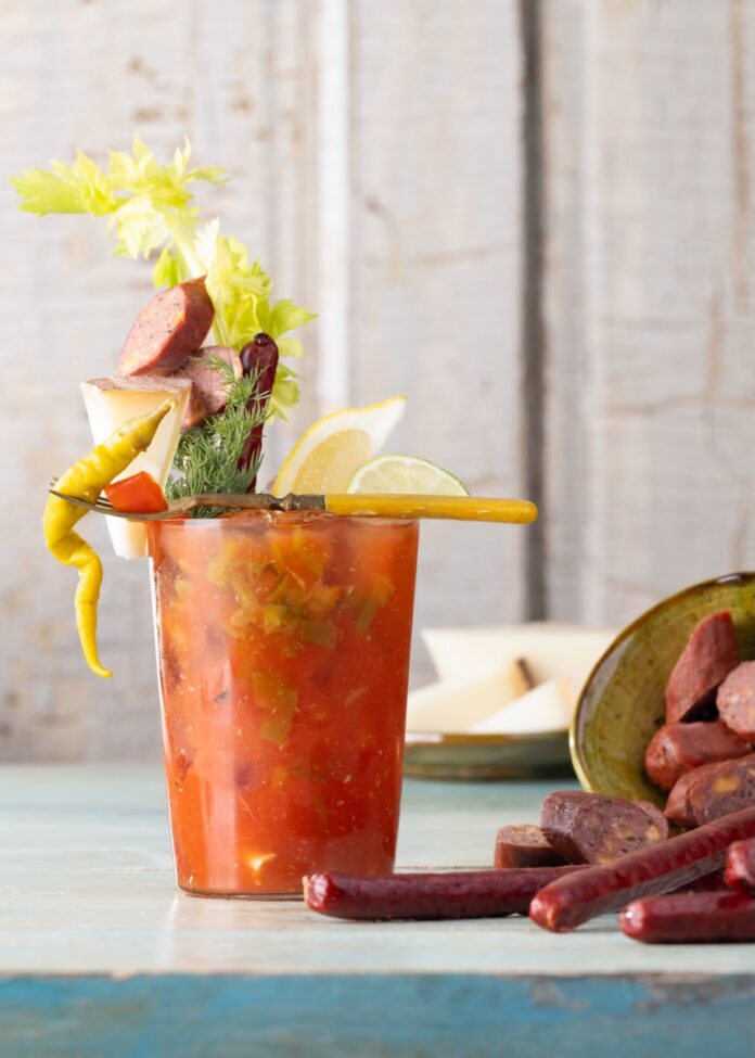A bloody mary sits in a glass on a table beside New Mexico sausages on the table and veggie garnishes on the drink.