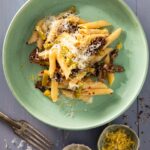 A light green bowl full of penne pasta with cheese and asparagus as a fork sits below the bowl.