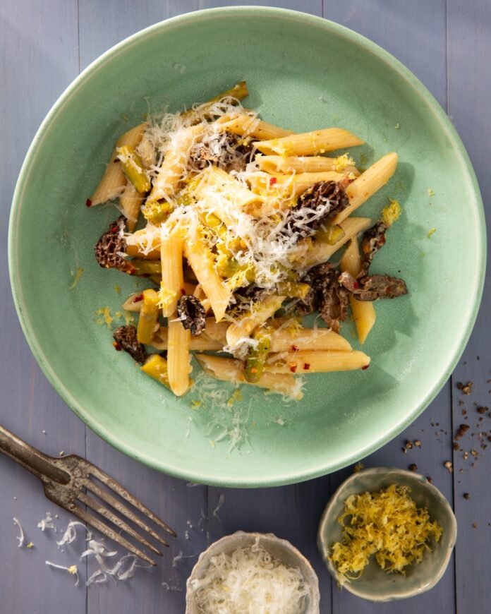 A light green bowl full of penne pasta with cheese and asparagus as a fork sits below the bowl.
