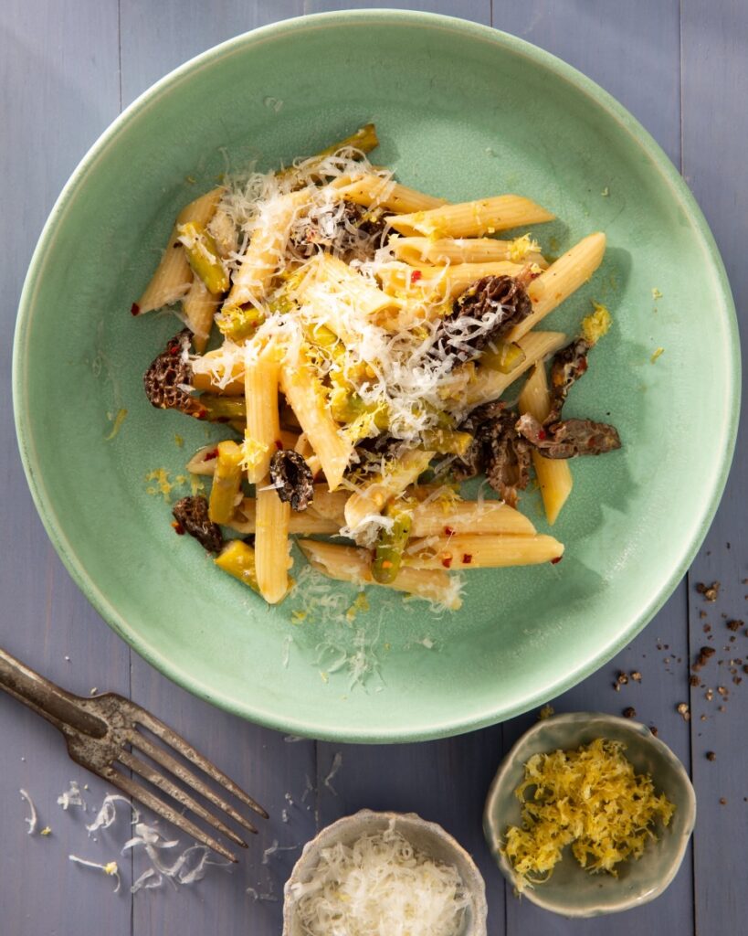A light green bowl full of penne pasta with cheese and asparagus as a fork sits below the bowl.