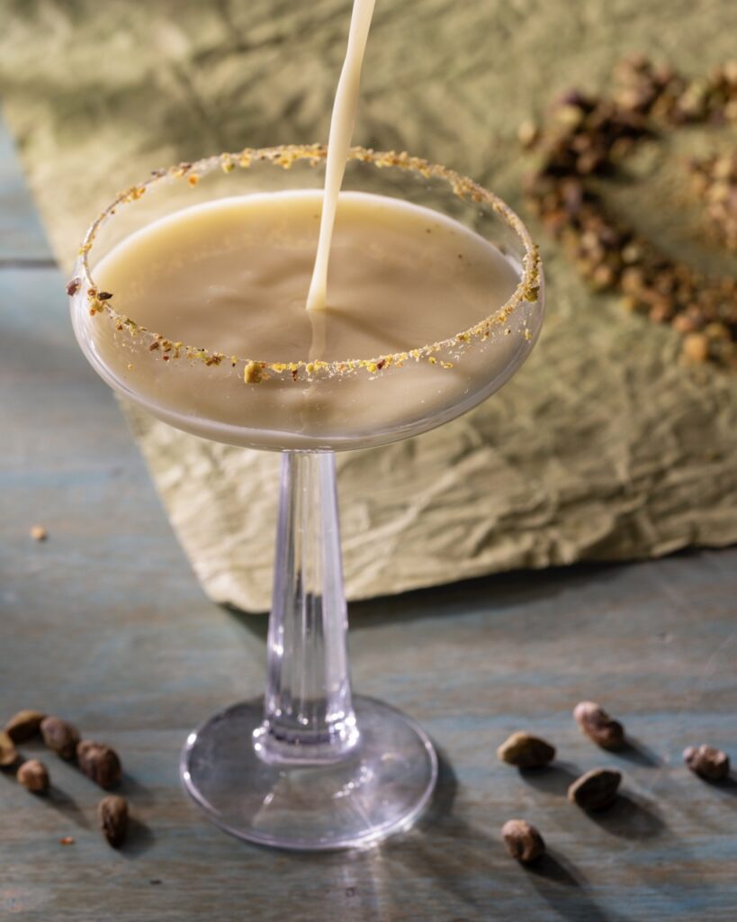 A Pistachio Martini gets poured into a coupe glass with nuts around the table.
