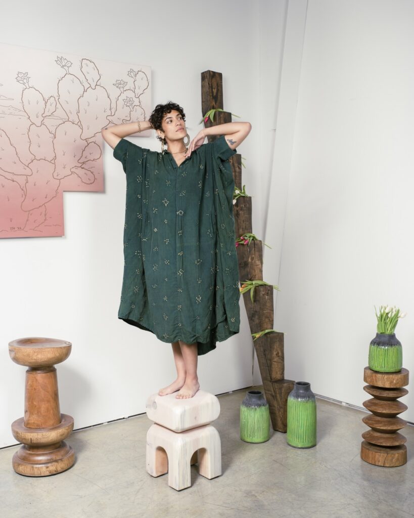 A woman stands on a stool in a long green dress with her arms up.