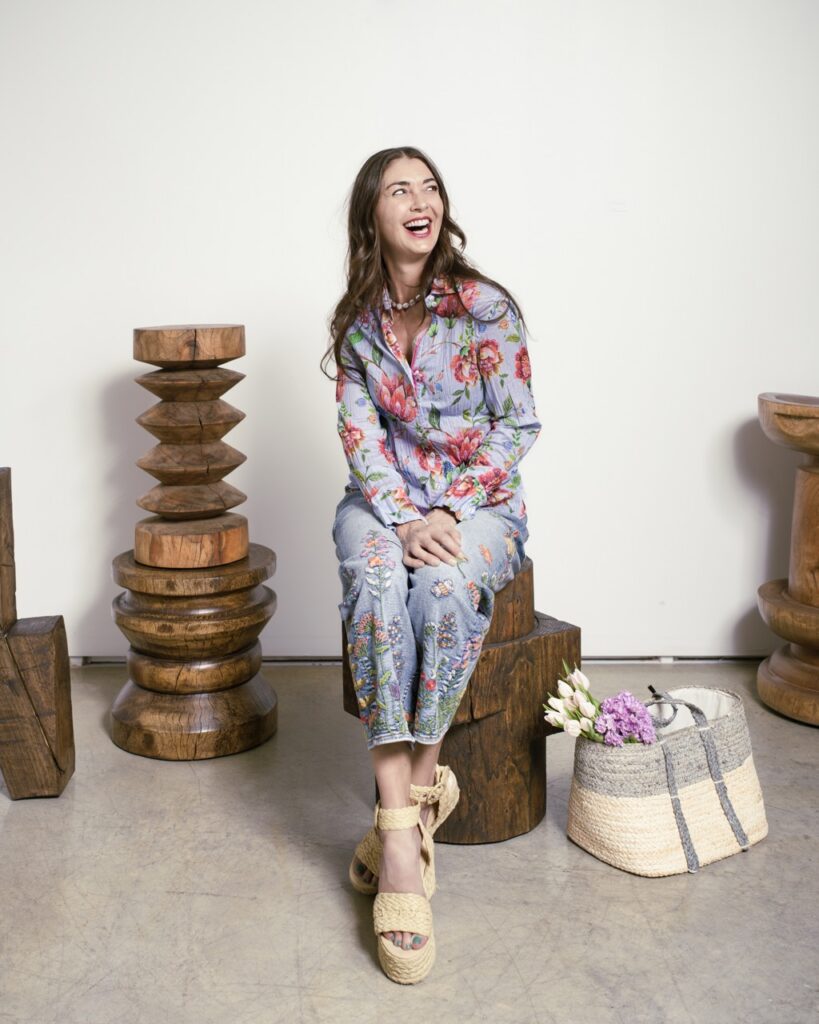 A woman sits on a stump in a studio with a floral shirt on.