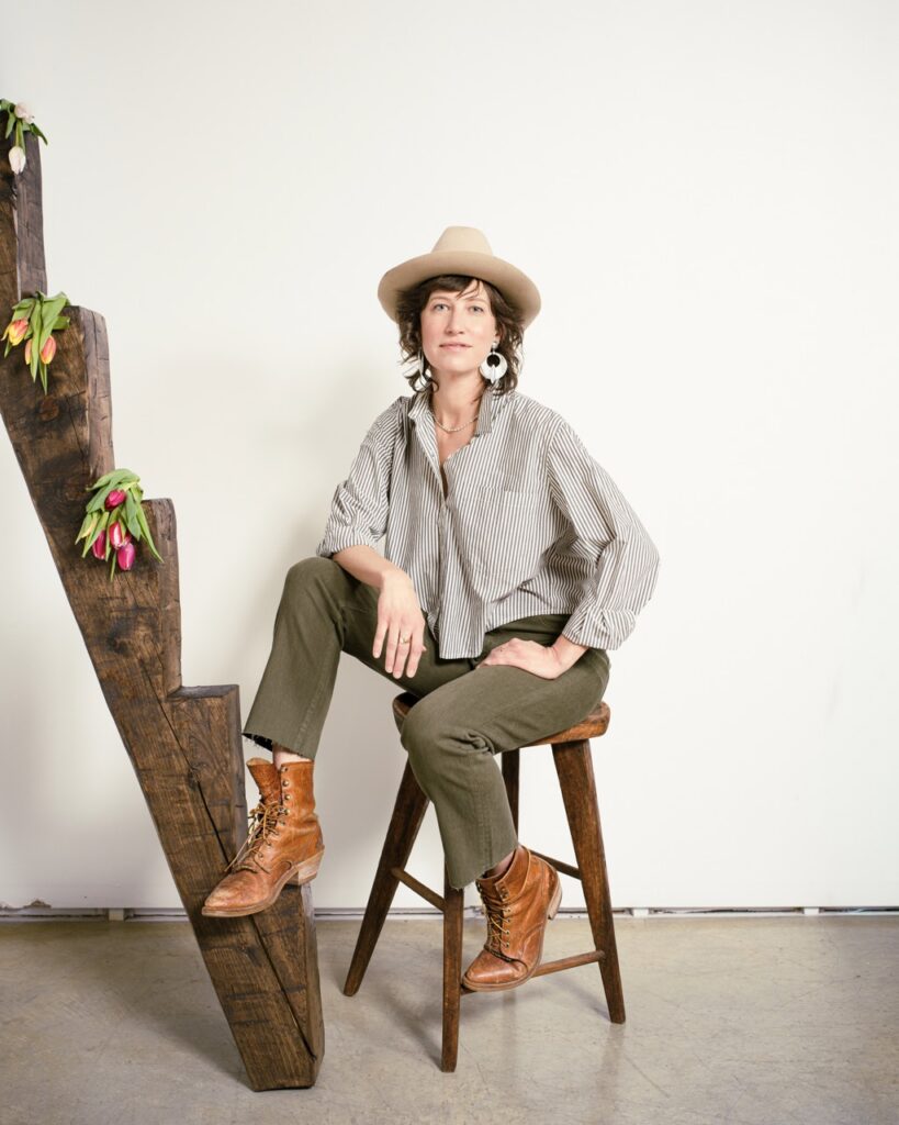 A woman in a grey jacket and green pants with a hat on sitting on a stool.