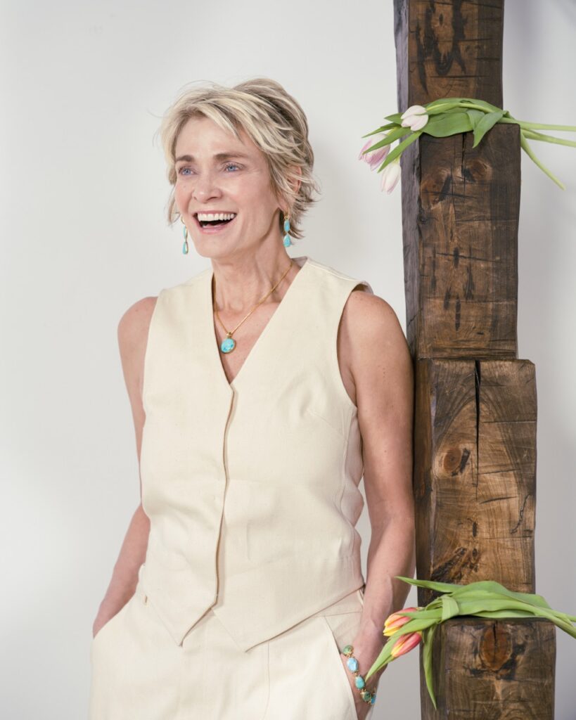 A woman laughs in a white vest and blue necklace.