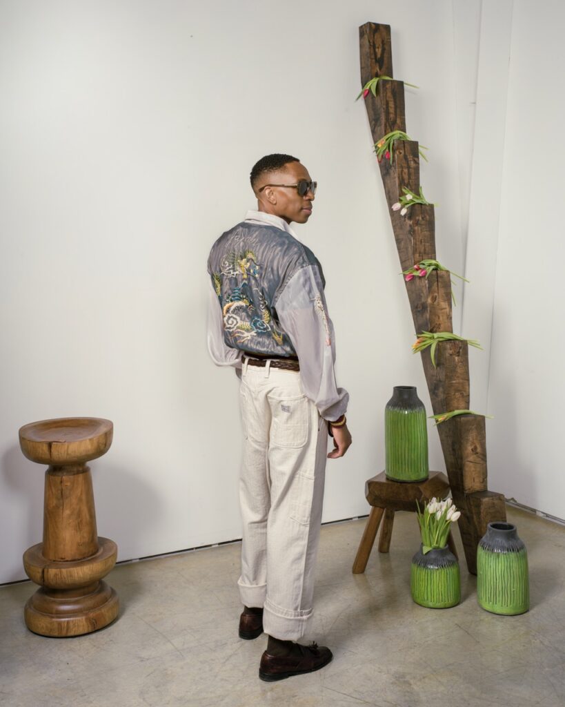 A man stands in a metallic shirt and white jeans with sunglasses on.