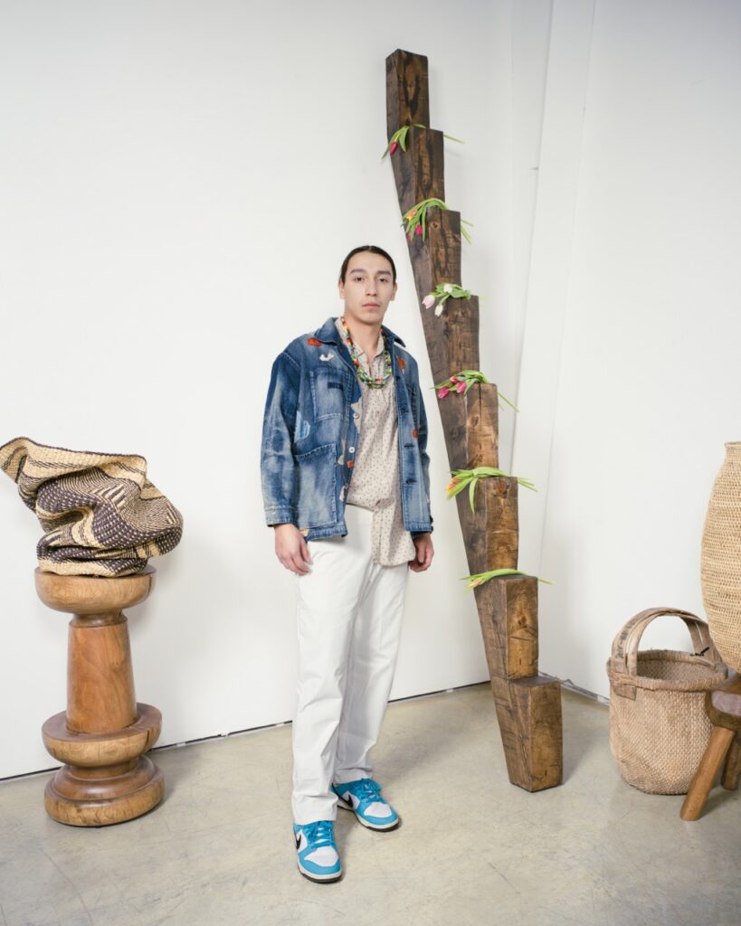 A man stands in a jean jacket, shirt, and white jeans.