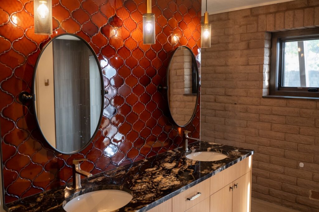 A dark orange tile bathroom wall with two circle mirrors and two sinks.