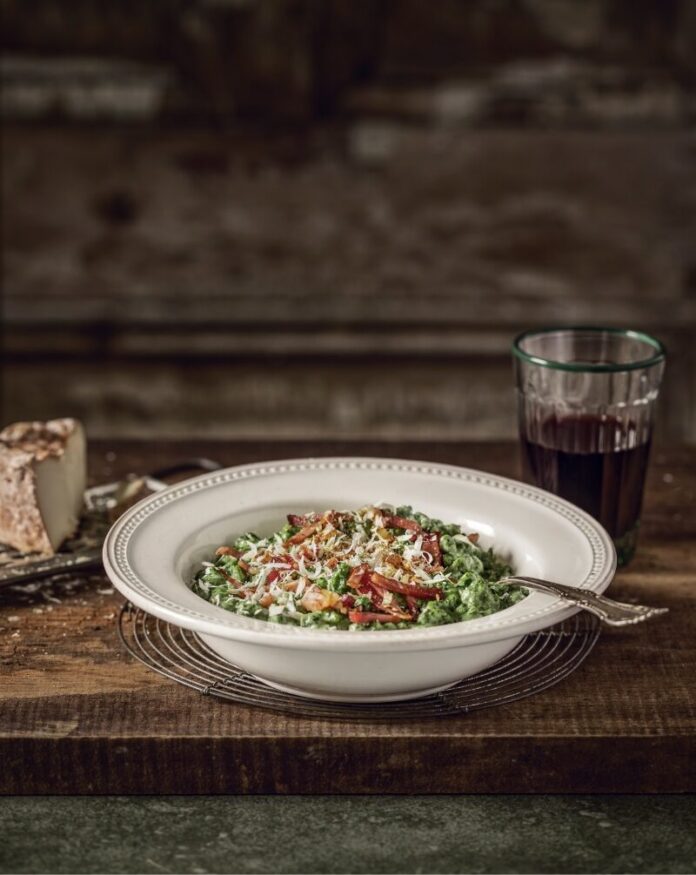 A deep white bowl of green spinach spätzle topped with crispy speck and grated cheese, next to a glass of red wine and a piece of cheese on a rustic wooden table.
