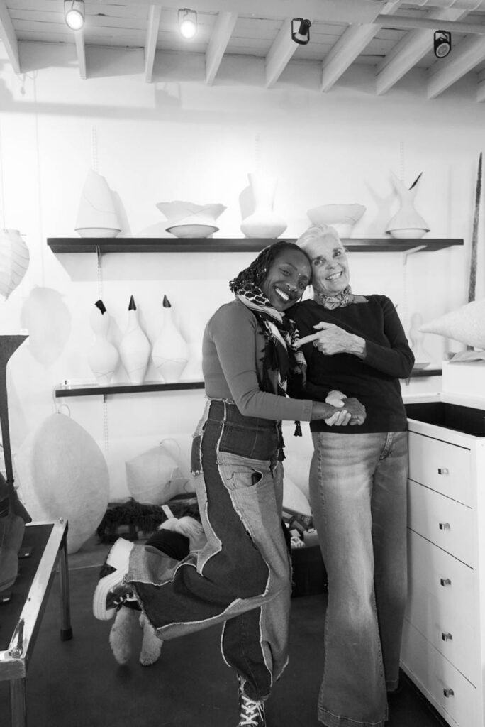 Two women embrace beside a desk with pottery behind them.