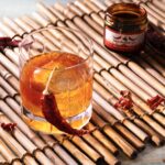 An old fashioned cocktail on a wood mat with a dried red chile hanging off it and a jar of chiles in the background.