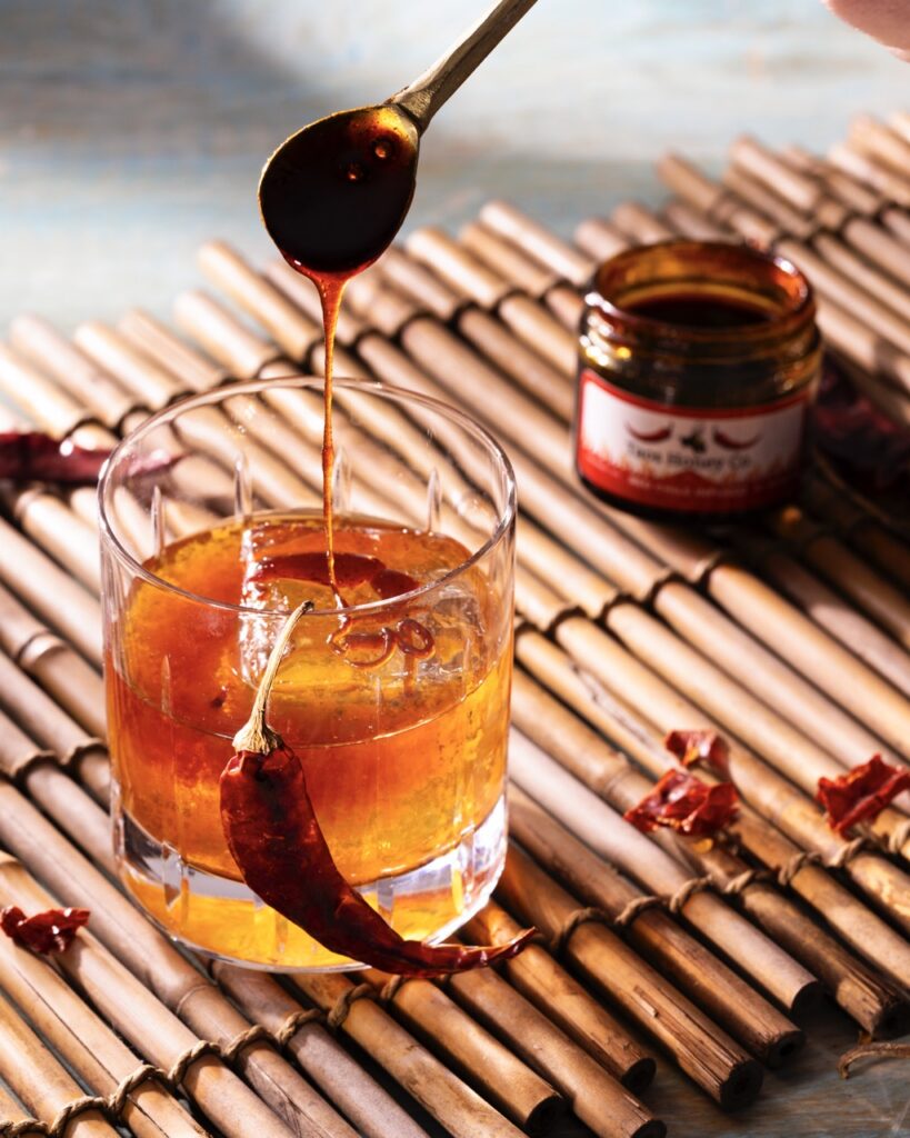 A spoon pours hot honey into an old fashioned cocktail with a dried red chile hanging off the side and a jar in the back.