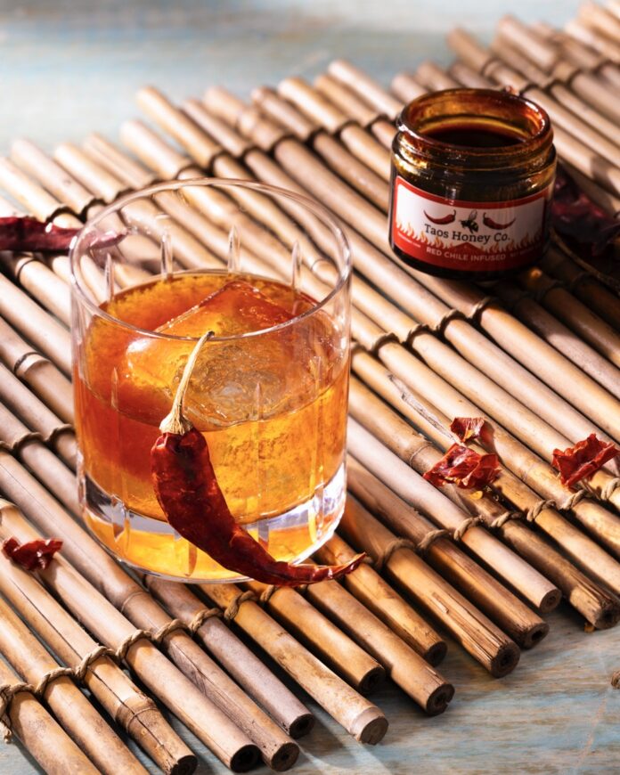 An old fashioned cocktail on a wood mat with a dried red chile hanging off it and a jar of chiles in the background.