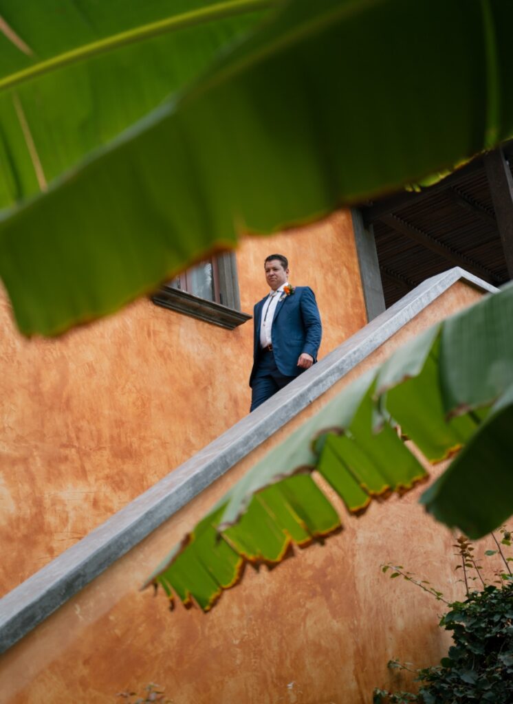 A groom in a light blue suit and orange boutonniere walking down an outdoor concrete staircase framed by large tropical green leaves.