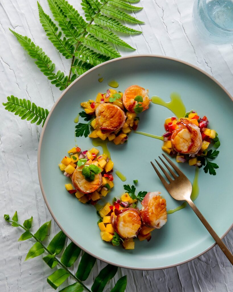 Four bacon wrapped scallops with fruity mango pineapple salsa sits on a blue plate.
