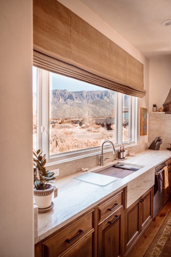 A rustic kitchen featuring custom wood cabinetry by Ernest Thompson and a neutral fabric window shade, overlooking the Sandia Mountains of New Mexico.