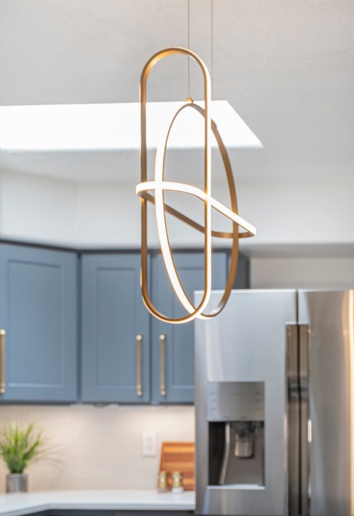 A modern kitchen with blue cabinetry by Hale Cabinets and stainless steel appliances from Builders Source, featuring a unique gold interlocking loop pendant light.