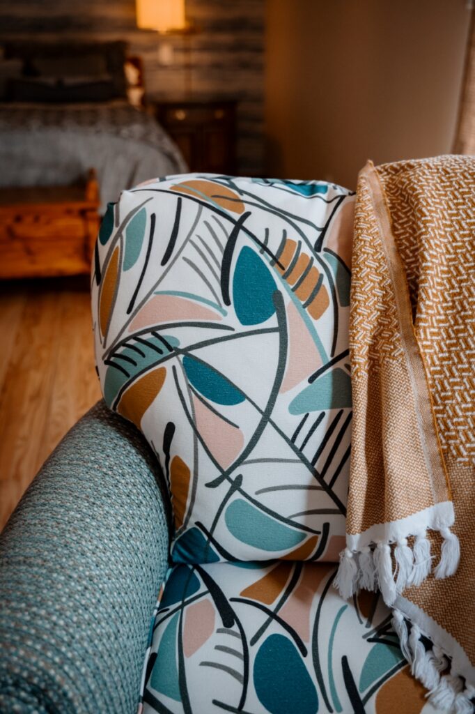 A decorative armchair featuring abstract leaf-patterned fabric from Fabricut and Norbar Fabrics, accented with a mustard yellow fringed throw, designed by Spatial Harmony.