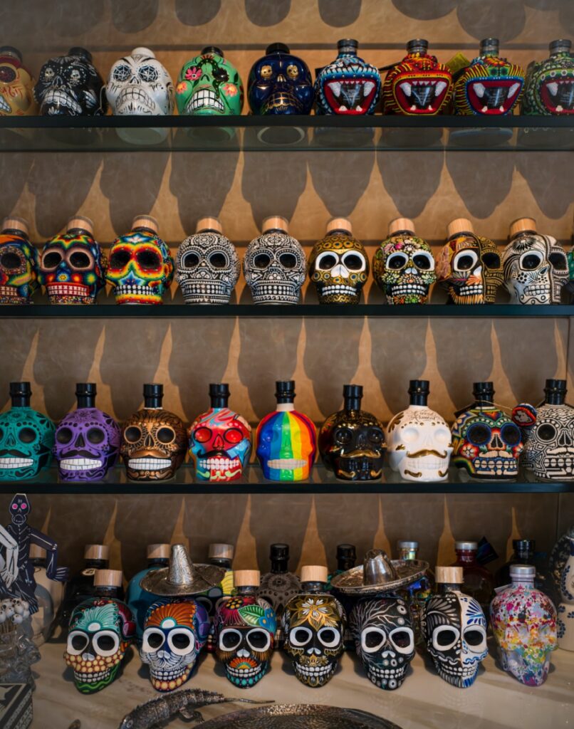A collection of over 50 artisan-made mezcal skull bottles, intricately hand-painted and beaded, displayed on glass shelves in David Arment and Jim Rimelspach’s Santa Fe home.
