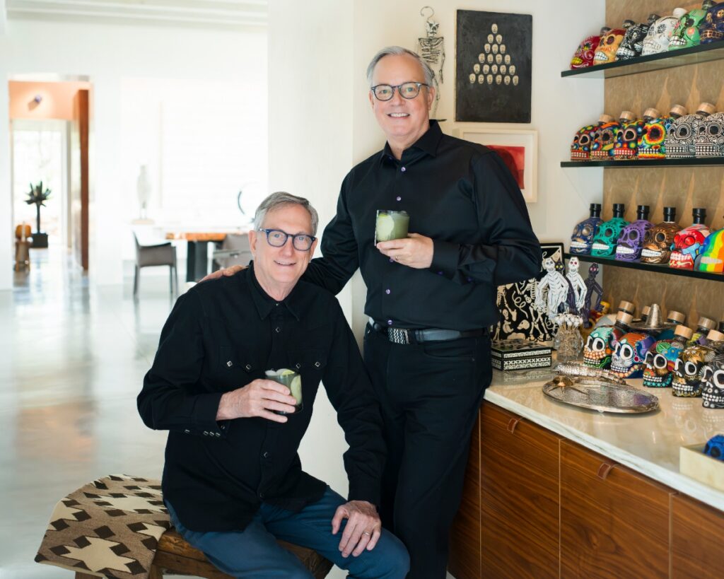 David Arment and Jim Rimelspach standing in their Santa Fe home next to a bar display of hand-painted Mexican mezcal skull bottles.