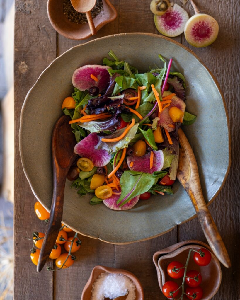 A vibrant garden salad in a rustic wooden bowl, featuring leafy greens, thinly-sliced watermelon radishes, and yellow cherry tomatoes served at an outdoor Santa Fe brunch.