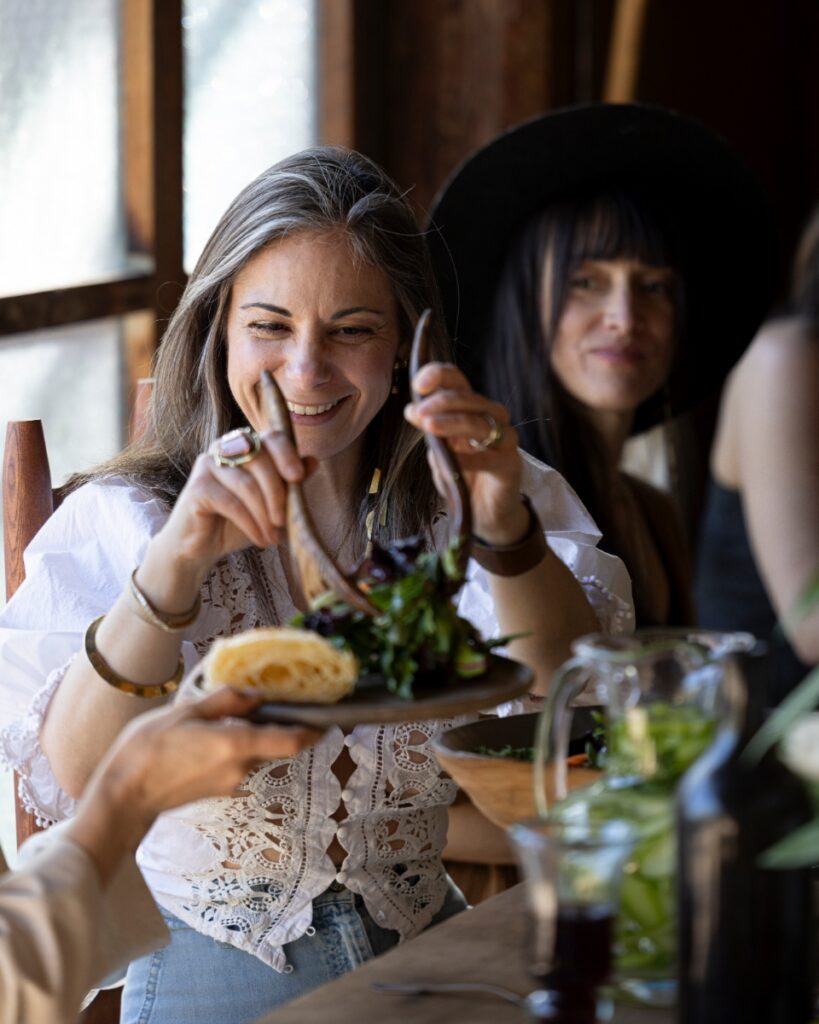 A woman smiling while serving a fresh green salad with wooden tongs during a casual outdoor house party in Santa Fe.