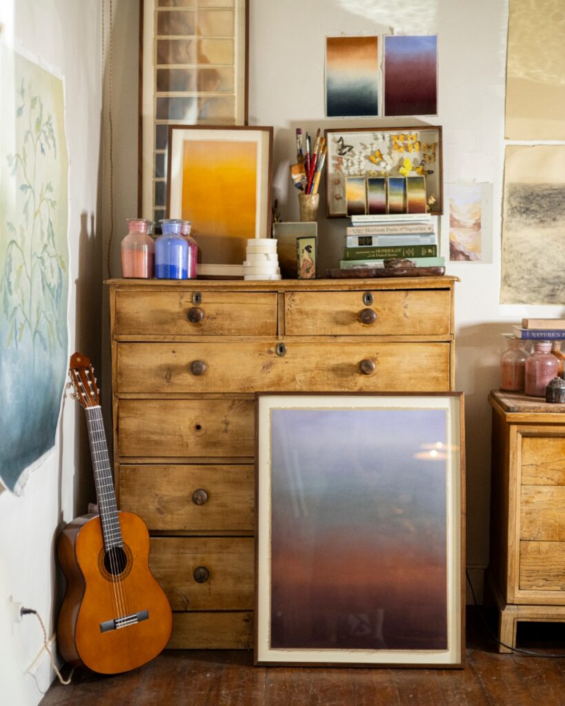 An artist's studio nook in a Santa Fe home featuring a vintage wooden dresser topped with pastel pigments, framed color gradient art, a butterfly display, and an acoustic guitar.