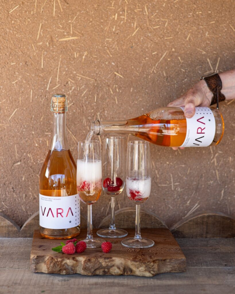 A person pouring Vara Silverhead Brut Rosado into champagne flutes filled with fresh raspberries on a rustic wooden board against an adobe wall in Santa Fe.