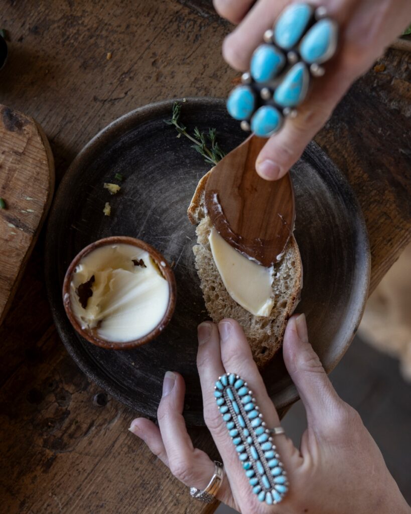 A person wearing turquoise rings spreading butter onto a slice of sourdough bread at a rustic Santa Fe dinner party.