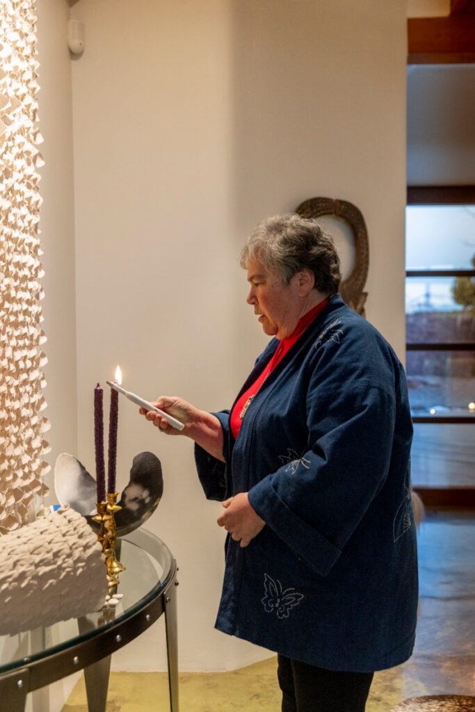 A woman in a blue butterfly-patterned jacket lighting purple candles for a Passover Seder ritual in a modern Santa Fe home.