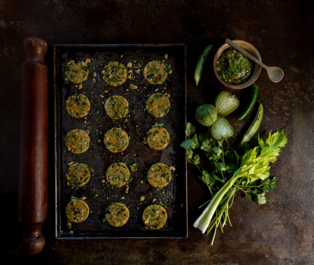 A tray of round, herb-flecked Passover crackers alongside fresh tomatillos, cilantro, and serrano peppers, reflecting a Mexican-inspired Sephardic Seder.