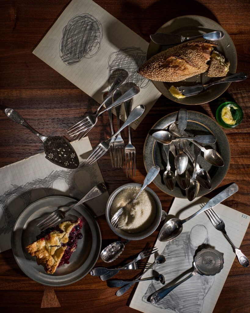 Patrick Mehaffy’s antique American coin silver cutlery collection arranged on a wooden table with sketches, bread, and a slice of berry pie.