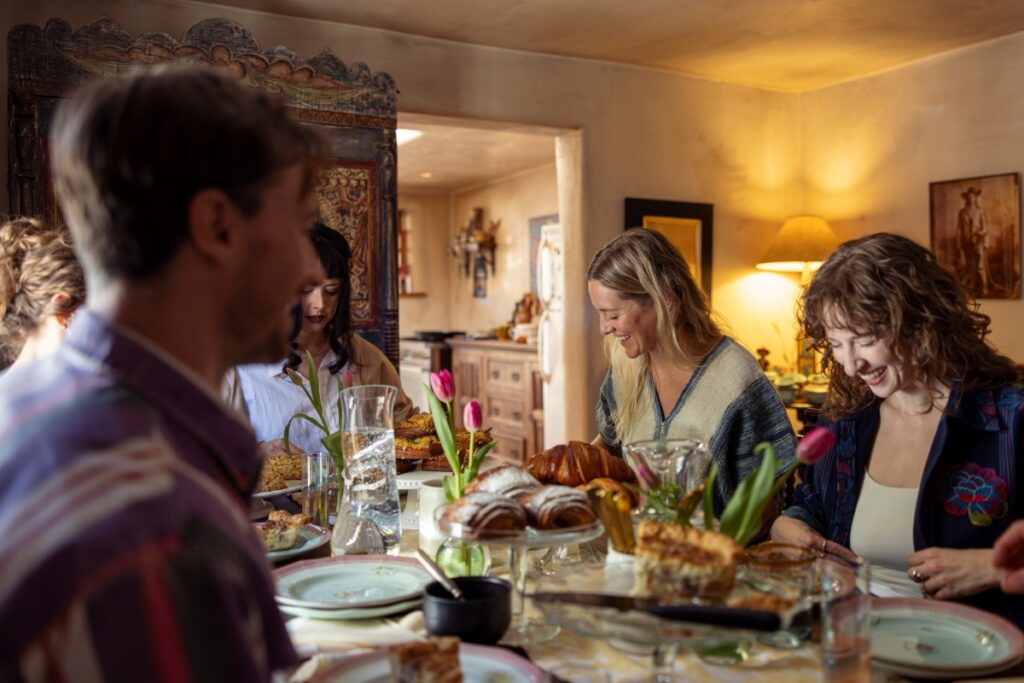 A candid moment of laughter between friends during an afternoon tea at a historic Santa Fe home, featuring a table filled with croissants and quiche.