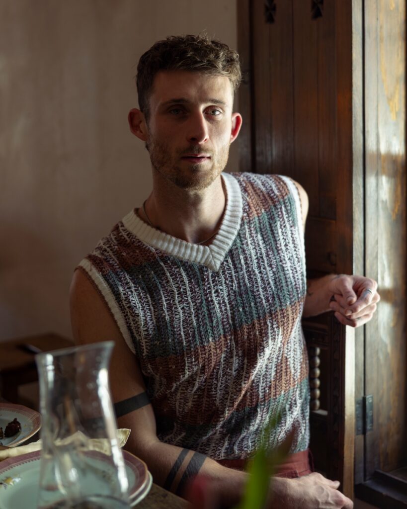 Pastry chef Chase Maus wearing a patterned sweater vest, sitting at a sunlit wooden table in a historic Santa Fe home.