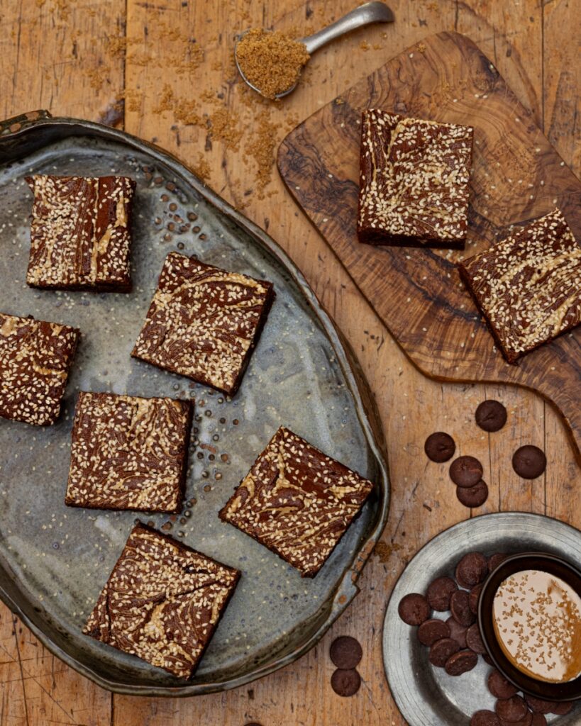 A flat lay of homemade chocolate tahini brownies topped with sesame seeds, served on a rustic metal tray and a wooden cutting board.