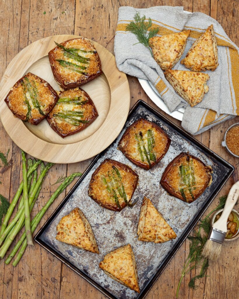 A rustic wooden table featuring freshly baked asparagus tarts on a baking sheet and savory herb scones wrapped in a linen cloth.