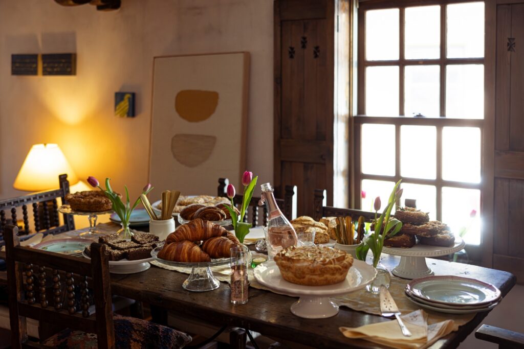 A festive afternoon tea spread on a wooden table in a historic Santa Fe home, featuring croissants, meringue pie, brownies, and pink tulips.
