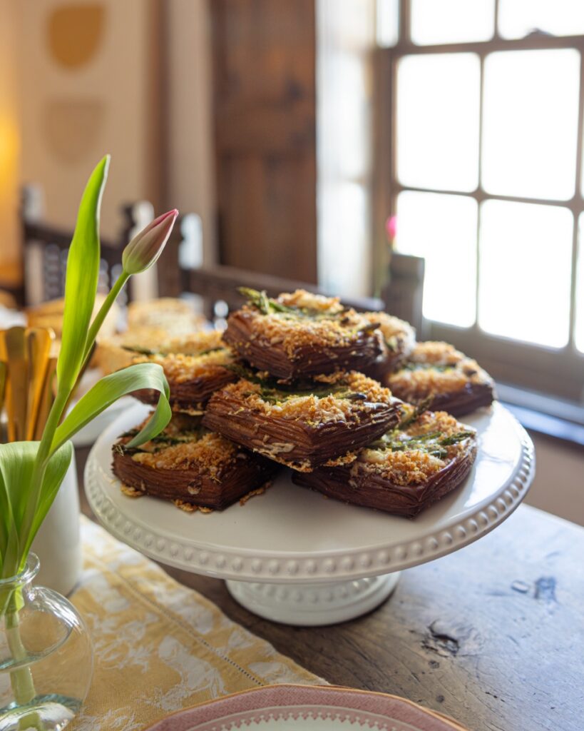 A stack of golden-brown asparagus and Gruyère puff pastry tarts served on a white beaded cake stand.