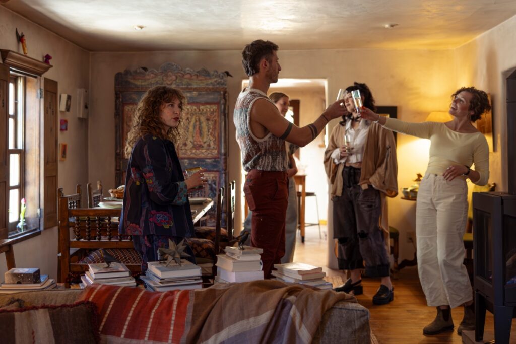A group of friends gathered in a sunlit, artistically decorated Santa Fe home, raising glasses for a toast near a table spread with fresh pastries.