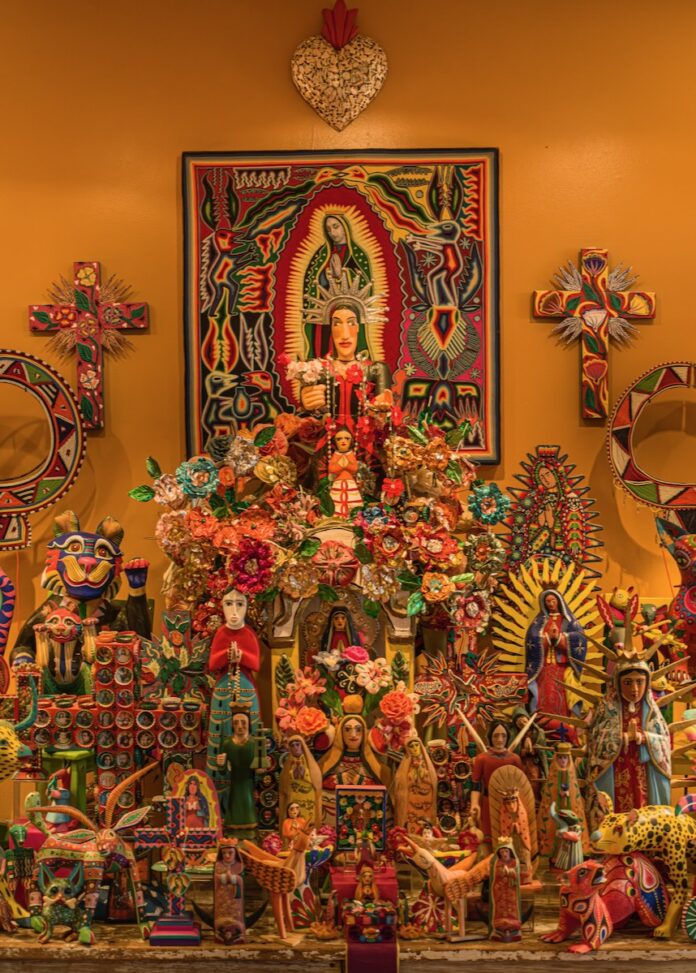 A dense and colorful display of Mexican folk art, including numerous hand-carved alebrijes wood sculptures, religious icons, and decorative crosses against a golden wall.