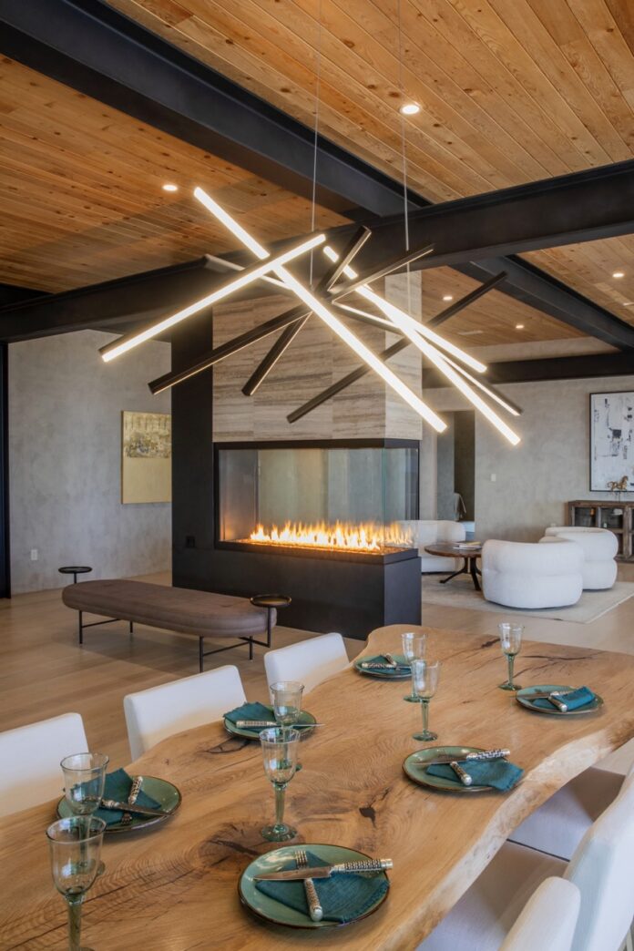 A fine pit in the middle of the room lights up while a wood dining table sits in the foreground and abstract light bars hang from the ceiling.