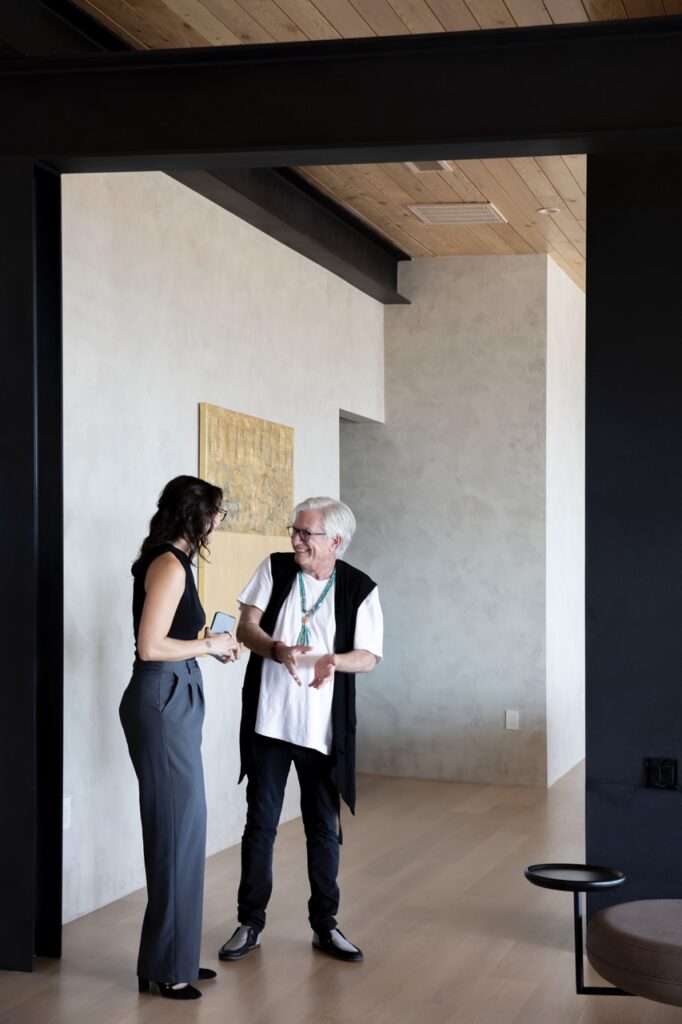 A woman in a white shirt and black vest talks to another in a black tank top while standing under a large black doorway.