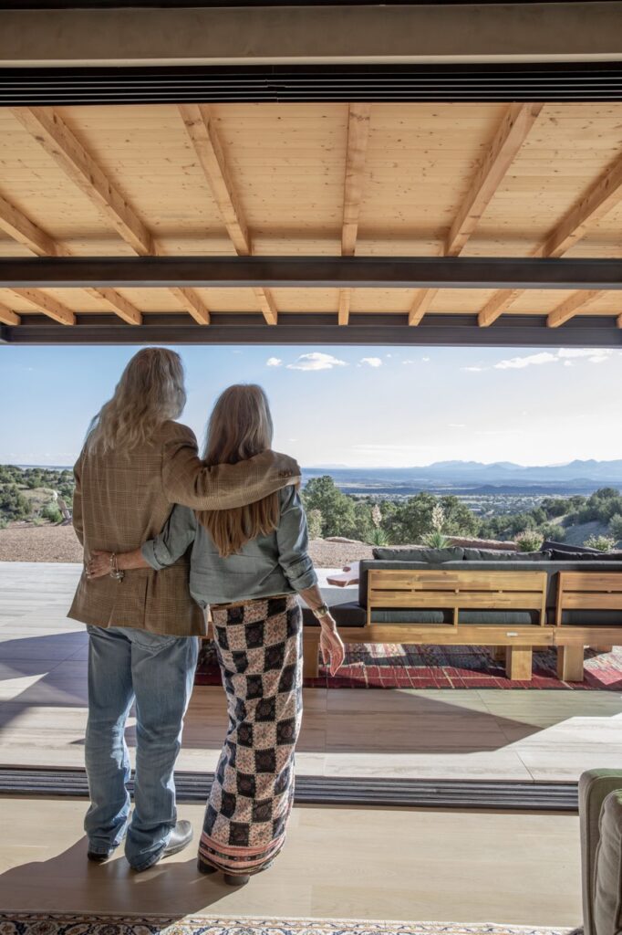 A man and woman stand arm in arm looking over a patio mountain top view of the desert.