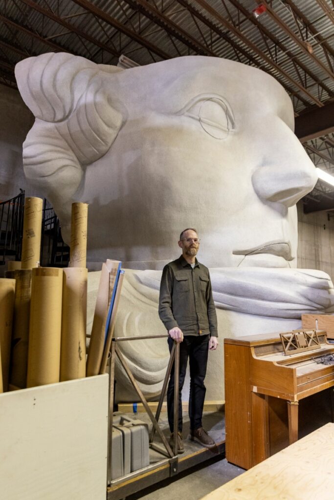 Director of Production Kris Longley-Postema (KLP) standing in front of a massive, white sculpted head set piece in the Santa Fe Opera scenic warehouse.