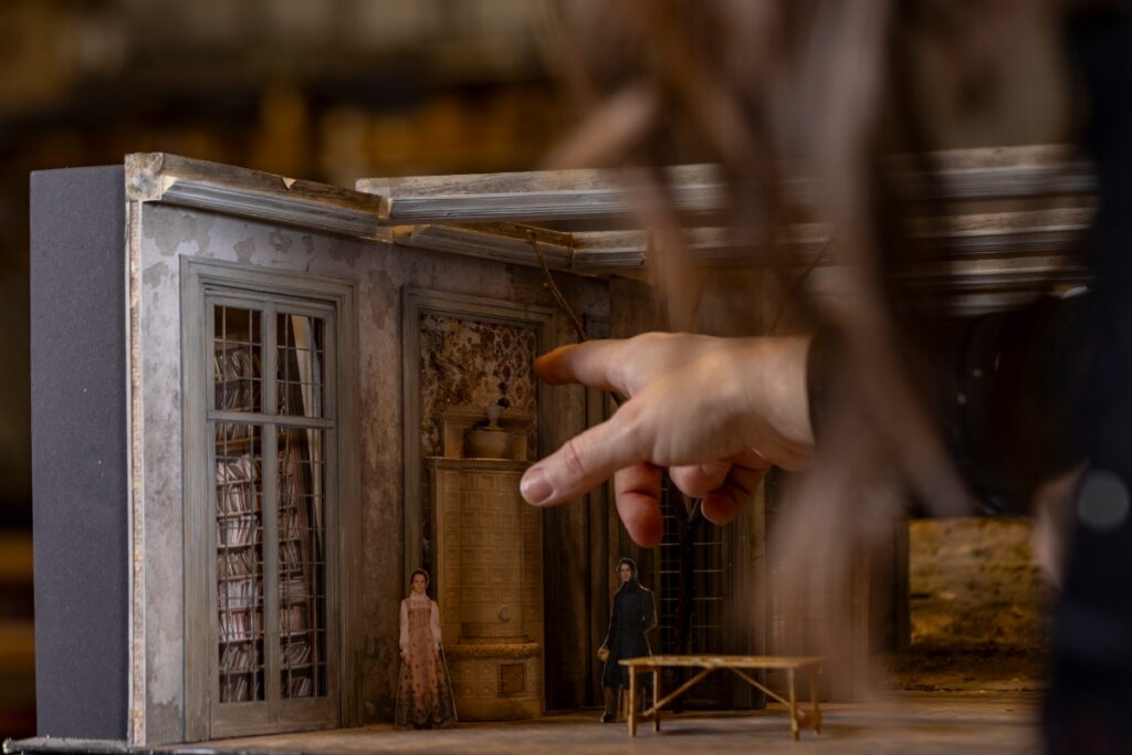 A detailed miniature set model for a Santa Fe Opera production, showing a classical room with tall windows, bookshelves, and a large ceramic heater.