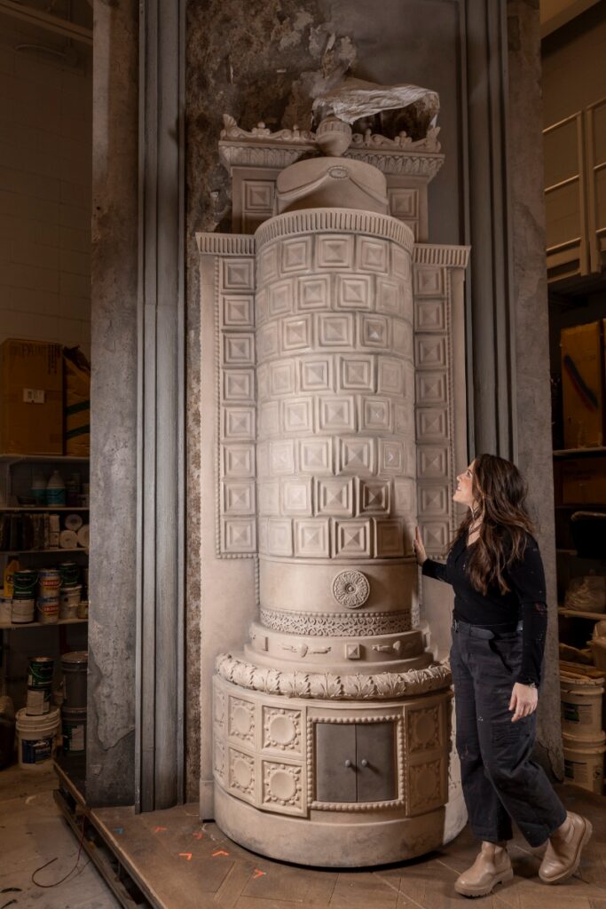 Scenic Charge Laura Greenfield standing next to a massive, intricately detailed white ceramic stove prop in the Santa Fe Opera scenic shop.