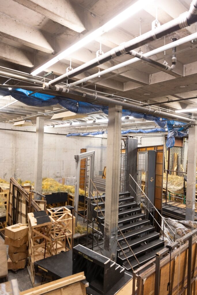A high-angle view of the Santa Fe Opera's expansive scenic warehouse, showing various stage sets, staircases, and production equipment in storage.