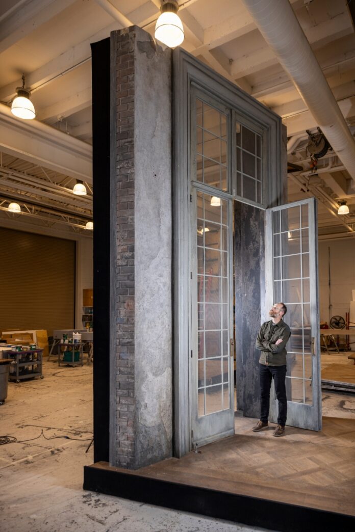 Director of Production Kris Longley-Postema (KLP) standing beside a massive, weathered classical door set piece in the Santa Fe Opera scenic shop.